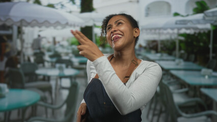 Woman pointing fingers in playful gesture showing hand and fingers, wearing apron by outdoor restaurant terrace tables and umbrellas; joy hospitality.