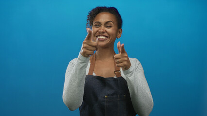 Woman pointing both index fingers at camera in blue studio wearing denim apron and gray long sleeve top; playful confidence.