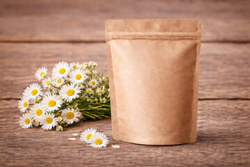 Blank kraft stand-up pouch mockup with chamomile flowers on rustic wooden table. Minimal eco packaging branding template, label design for organic herbal tea, copy space.