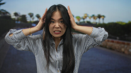 Woman outdoors feeling frustrated, wearing striped shirt, holding head in hands, expressing stress...
