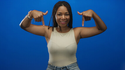 Woman pointing down with fingers at chest and hands visible in blue studio; confidence invitation welcome.