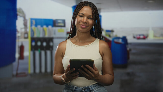 Woman holding tablet with hands visible at a petrol station on street, smiling while facing camera and standing near pumps; confidence focus. - Powered by Adobe