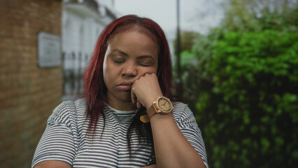 Woman rests cheek on hand wearing wooden watch in front of brick building beside hedge under...