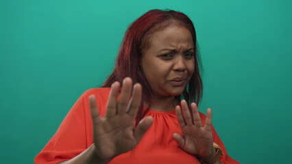 Woman holds up hands in a stop gesture with a scrunched face wearing an orange top in green studio;...