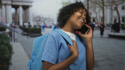 Woman in blue shirt smiling while holding backpack strap talking on phone on street; joyful connection.