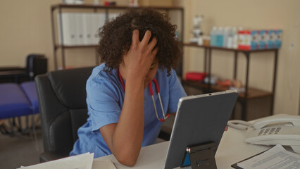Woman wearing blue scrubs and stethoscope holding forehead and reading tablet at medical desk in...