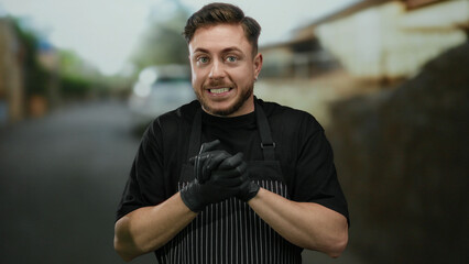 Young caucasian man with beard and apron wearing gloves, stands on a city street outdoors, appearing as a waiter.