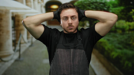 Fototapeta premium Young man with beard wearing apron and gloves stands outdoors on city street looking pensive with arms behind head, capturing a candid moment of urban life.