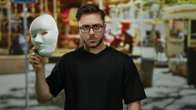 Young bearded man at outdoor fair removing white mask with blurred park background displaying casual attire and facial expression shift.