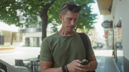 Man typing on smartphone with hands on screen on street under tree, wearing green tshirt and sunglasses; focus everyday life.
