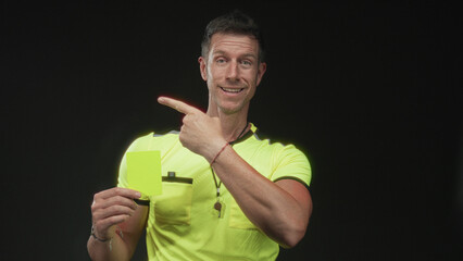 Referee man pointing finger and holding yellow card in studio with black background, wearing neon yellow shirt and whistle; authority discipline.