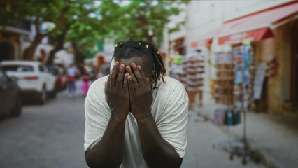 Fototapeta premium Young african american man in a white t shirt with hands covering his face on a crowded city street lined with storefronts and parked cars; embarrassment.