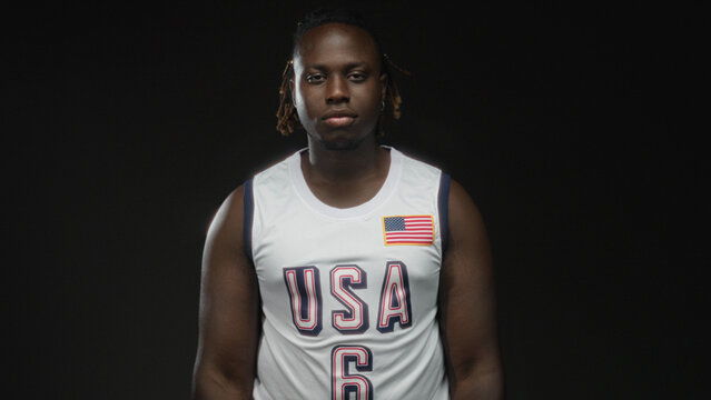 Young african american man in usa basketball jersey waving hand in black studio with direct gaze; quiet confidence.