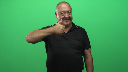 Fototapeta premium Senior hispanic man giving thumbs up with right hand extended wearing a black polo against a green studio backdrop; approval confidence warmth.