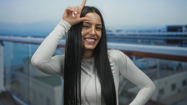 Woman gesturing loser sign on cruise boat with ocean in background, wearing white shirt, smiling and enjoying outdoor seaside view on a sunny day, exuding cheerful and youthful energy.