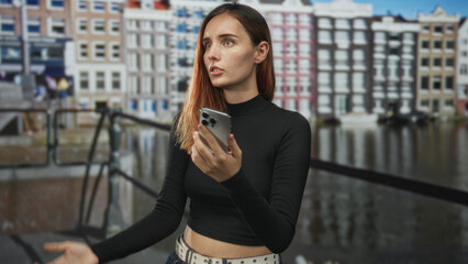 Redhead woman frowns at smartphone while holding it with both hands on a street by canal buildings; frustration isolation.