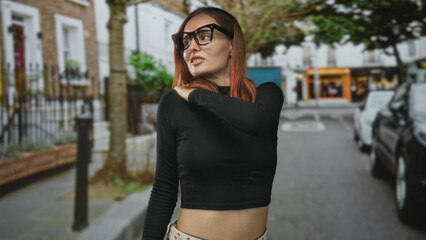 Young woman touching her shoulder and showing bare midriff, wearing oversized glasses on a street; pensive solitude.