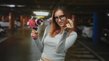 Woman holding red tape dispenser and showing pinched fingers gesture in parking building; playful mischief.