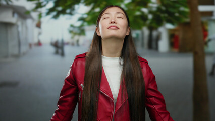 Woman in red jacket enjoying peaceful moment outdoors on a city street during daytime with gentle light and relaxed expression, surrounded by soft urban background and nature.