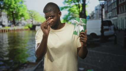 Young handsome man holding green gift box, pinching fingers near white ribbon bow while standing on street; skepticism curiosity.