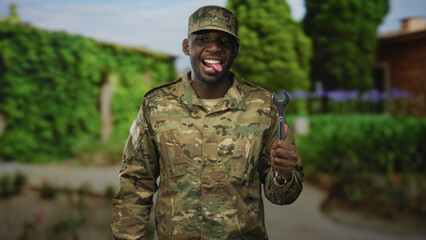 Man soldier in camouflage uniform holds wrench, sticks out tongue in front of building; pride duty resilience.