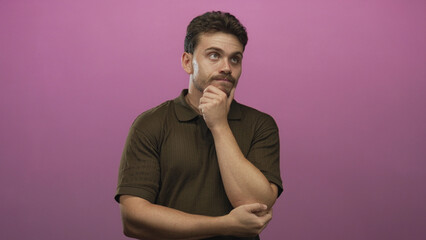 Man with hand on chin in pink studio, young hispanic displaying pensive pose and folded arm...