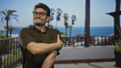 Naklejka premium Man in brown polo shirt holds and rubs bare forearm on building balcony overlooking palm trees and ocean; discomfort seaside scene.