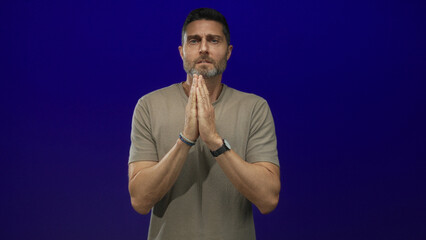 Man with clenched fists and bare hands held near chest in studio against deep blue backdrop; desperation plea.