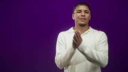 Obraz premium Young hispanic man claps hands and smiles, wearing white sweater and glasses in a purple studio; celebration joy.