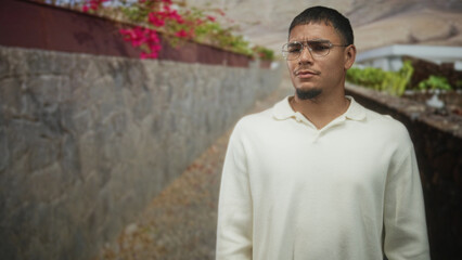 Obraz premium Man wearing glasses stands with hands at sides on a stone street beside a planter and wall, looking slightly to the right; contemplation.