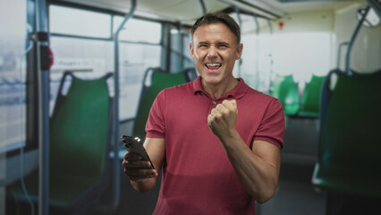 Man holding smartphone pumps clenched fist while seated on a bus on a city street, green seats visible; triumph.