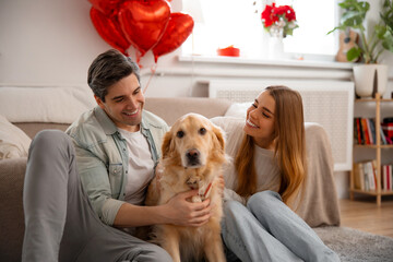 Smiling young couple relaxing on sofa with their dog at home while celebrating Valentine’s Day. Love, family, pets and cozy lifestyle indoors.