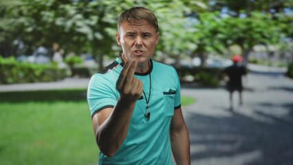 Man referee wearing teal tshirt with whistle on lanyard, beckons with raised hand and stern expression amid park trees on street pathway; authority discipline.