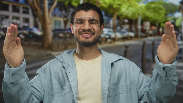 Man measures width with hands near face on busy street under green trees with parked cars behind; evaluation happiness.
