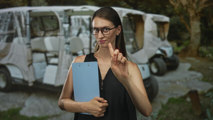 Woman holding blue clipboard with both hands in forest near parked golf carts, wearing black...