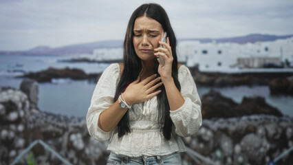 Woman holds phone to ear with hand on chest, wearing white blouse and jeans at building by rocky seaside with distant white houses; sorrow.