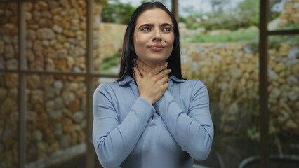 Woman indoors suffering throat pain holds neck appearing uncomfortable against stone wall in bright...