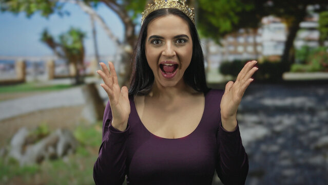 Young hispanic woman with crown expresses surprise in an outdoor park filled with trees and sunlight, enhancing her joyful demeanor and vibrant atmosphere.