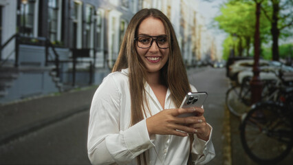 Fototapeta premium Woman holding smartphone and typing with both hands while smiling on a city street lined with bicycles and townhouse steps; happiness.