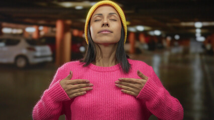Woman wearing pink sweater and yellow hat stands indoors in parking lot with cars in background,...