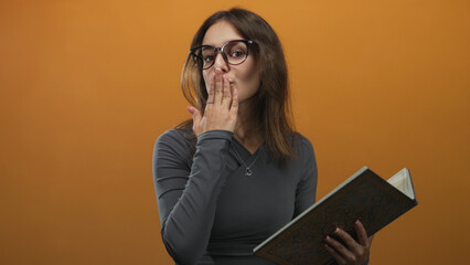 Young hispanic woman holding book blows kiss with hand to mouth in studio with orange backdrop; playful warmth.