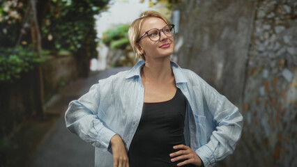 Blond woman wearing glasses in a light blue shirt and black tank, hands on hips and slight head...