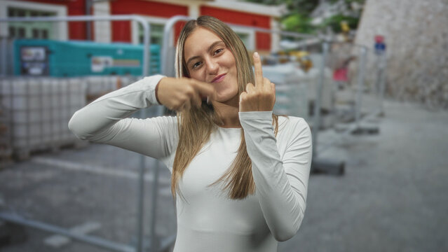 Woman in white long sleeve top pointing finger and showing middle finger near metal barriers on urban street; playful defiance.