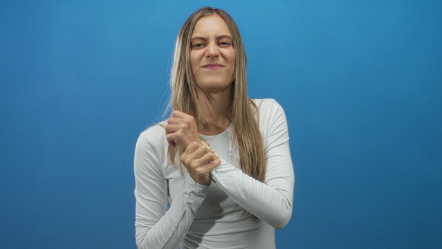 Woman clutching her bare wrist with both hands in a blue studio while wearing a white long sleeve top; discomfort recovery. - Powered by Adobe