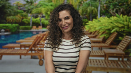 Naklejka premium Woman smiles with bare arms and curly hair while tilting head at resort poolside under bright sunlight; serenity.