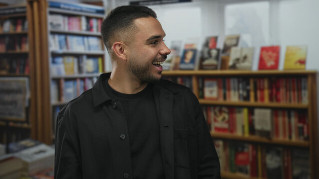 Young man wearing black jacket smiles and turns head near bookshelf while speaking in library; curiosity exploration.