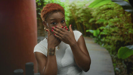 Young african american woman covering mouth with hands in forest wearing white top; surprise awe wonder reflection.