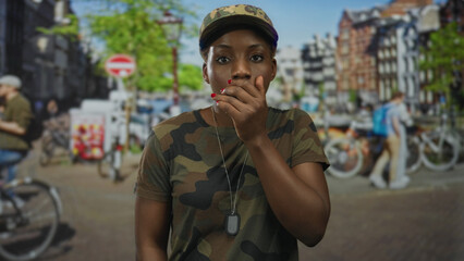 Young african american woman soldier in camouflage uniform covers mouth with hand on busy street;...
