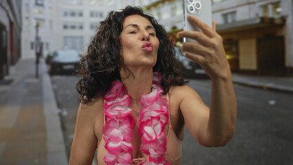 Fototapeta premium Middle age hispanic woman in pink bikini blows kiss to camera while holding smartphone on a busy city street; affection flirtation joy.