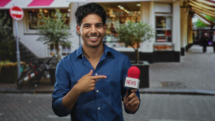 Man smiling and holding news microphone on street, wearing blue shirt with red mic foam and storefront in view; confidence warmth.
